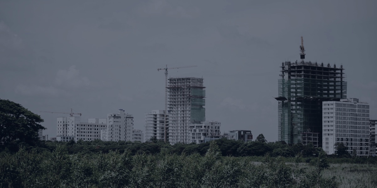 Prédios residenciais ao longe, alguns ainda em construção, com tela de proteção e guindastes, atrás de uma área de mato
